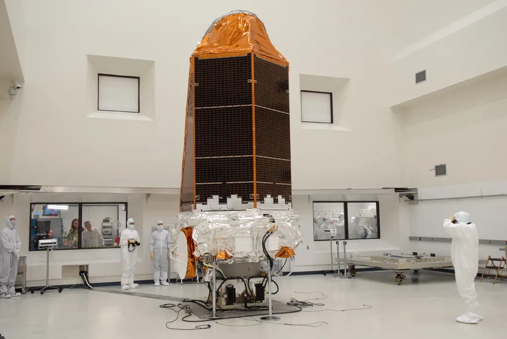 NASA's Kepler space telescope standing in a clean room before launch: a tall body wrapped in gold foil and dark solar panels, with technicians in white suits beside it for scale.